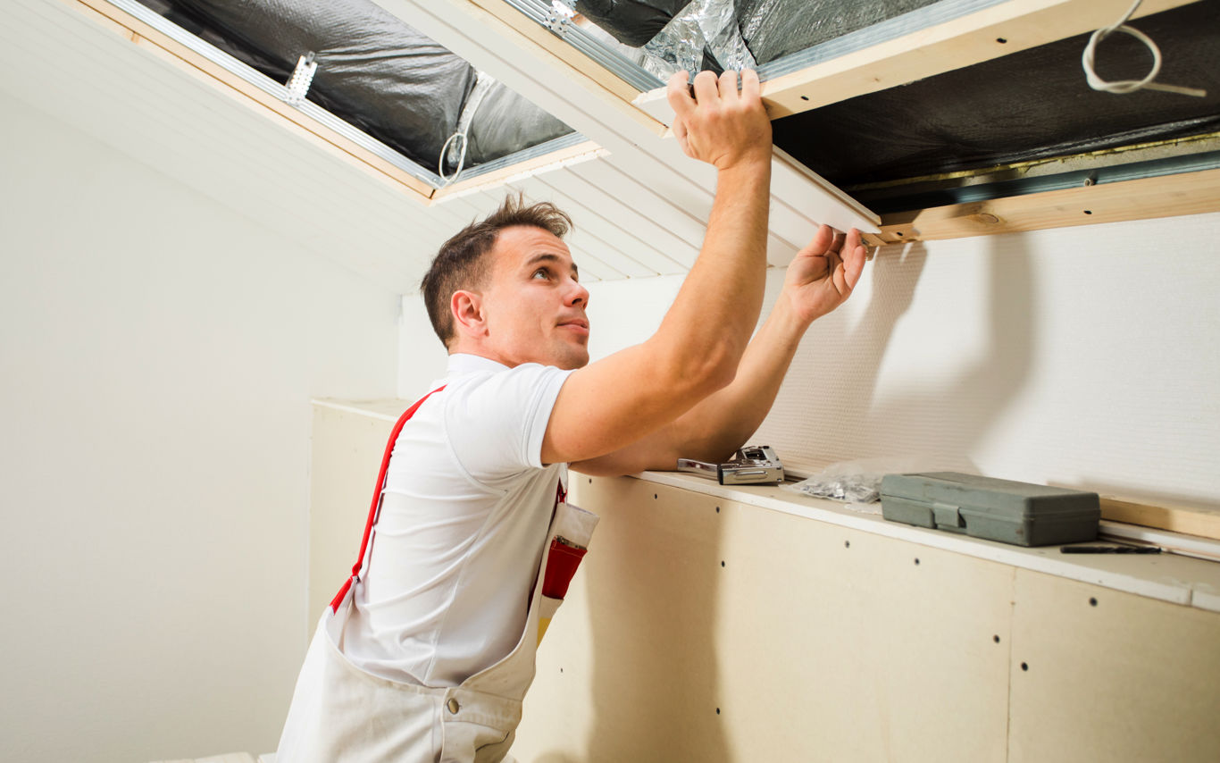 Håndværker iført hvid trøje og arbejdsbukser arbejder på montering eller reparation af et skråloft. Han bruger begge hænder til at justere en træramme, mens værktøj og materialer ligger på en arbejdsflade ved siden af.