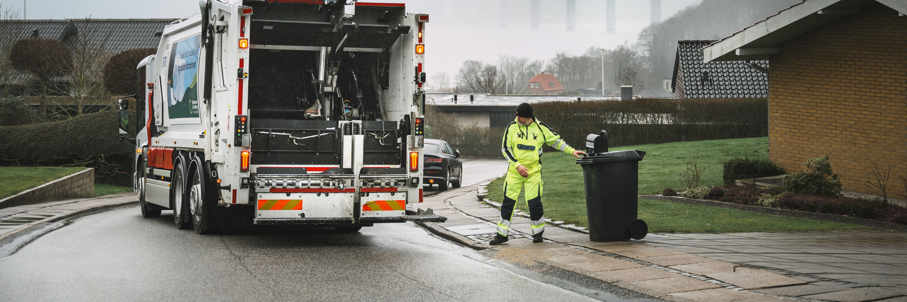 Bagenden af en skraldebil under arbejdet, med udsigt over et boligområde.